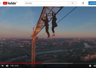 In einem YouTube-Video ist ein großer Baukran zu sehen. An seinem Ausleger in schwindelerregender Höhe hängen ungesichert zwei Personen, die sich nur mit den Händen festhalten.